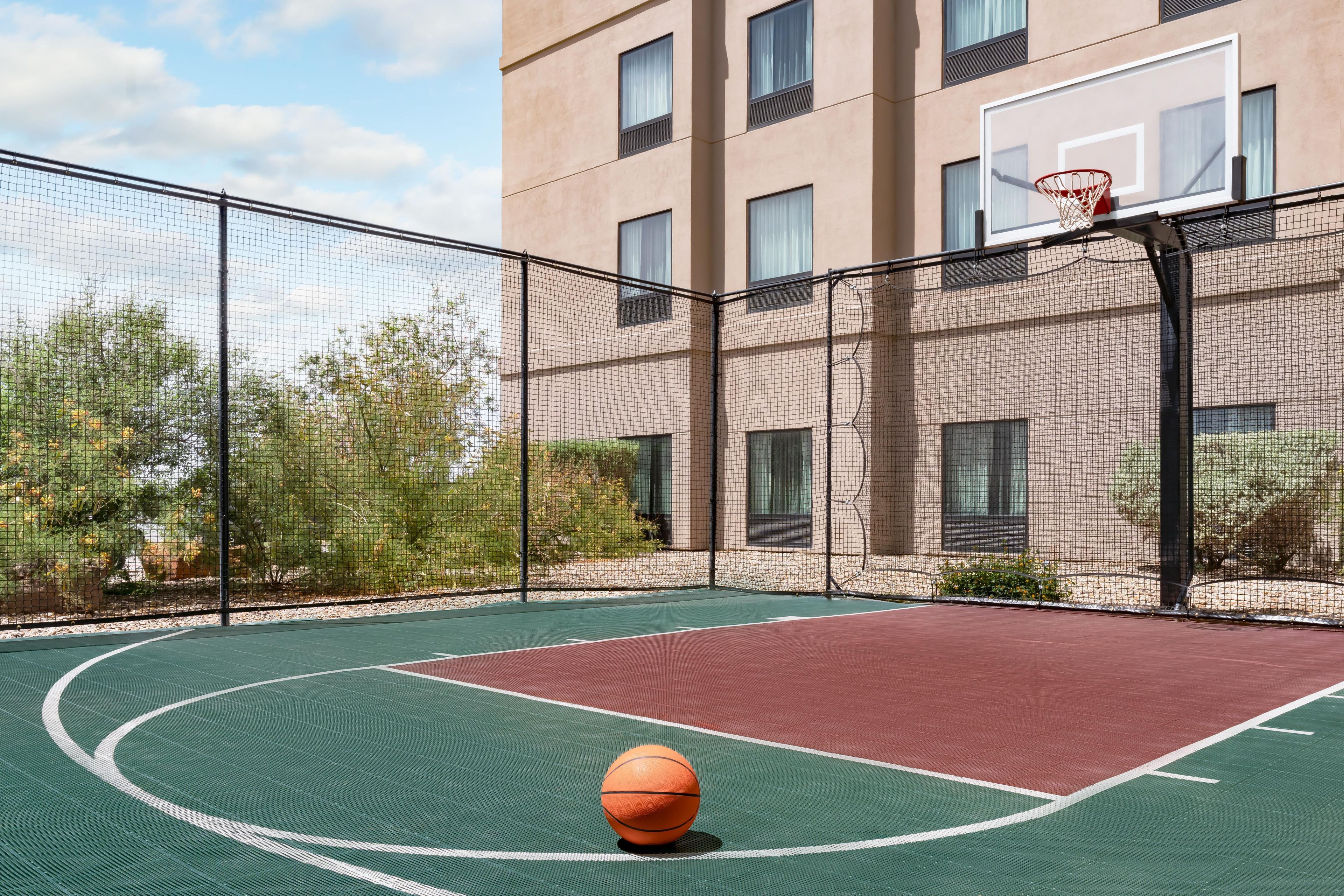 An outdoor basketball court with a hoop, surrounded by a fence, and a building in the background.