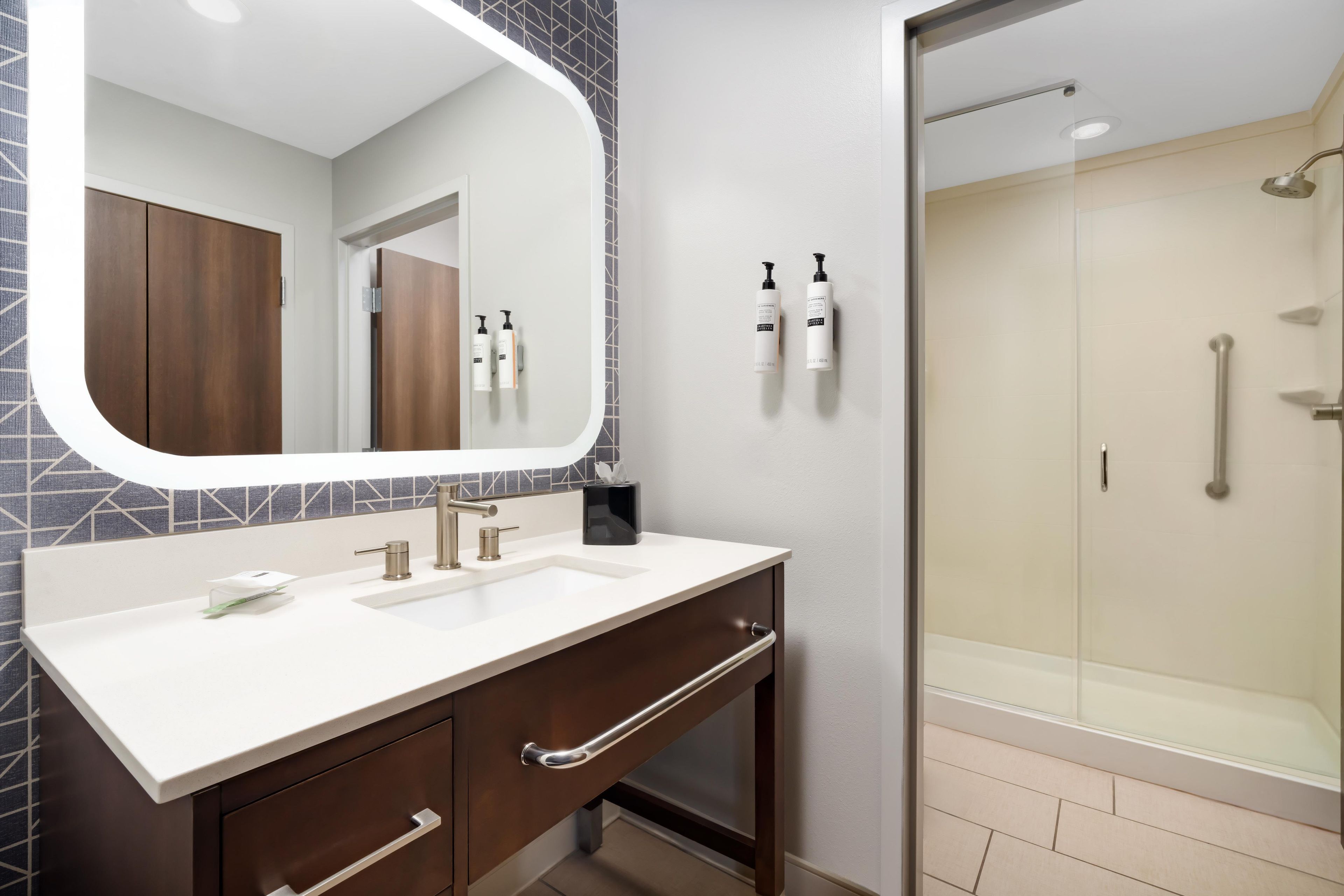 A bathroom with a large mirror, a sink, a shower, and toiletries on the wall.