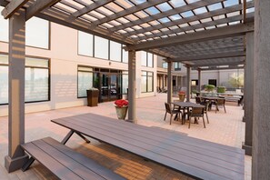 An outdoor patio area with wooden tables and benches, a pergola structure, and large windows.