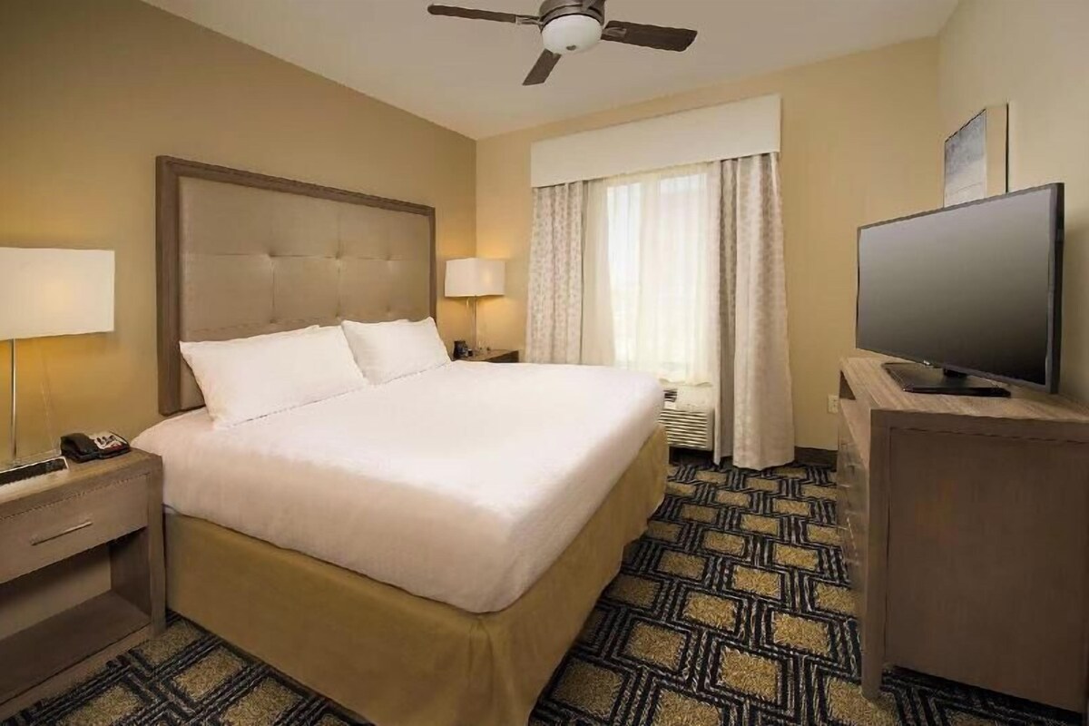 A hotel room with a bed, bedside lamps, a television, a ceiling fan, and a patterned carpet.