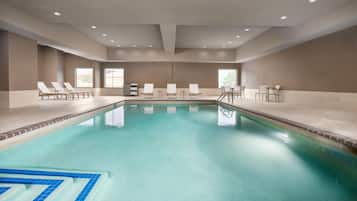 Una piscina interior con sillones de descanso y mesas, ventanas amplias y suelo de baldosas.