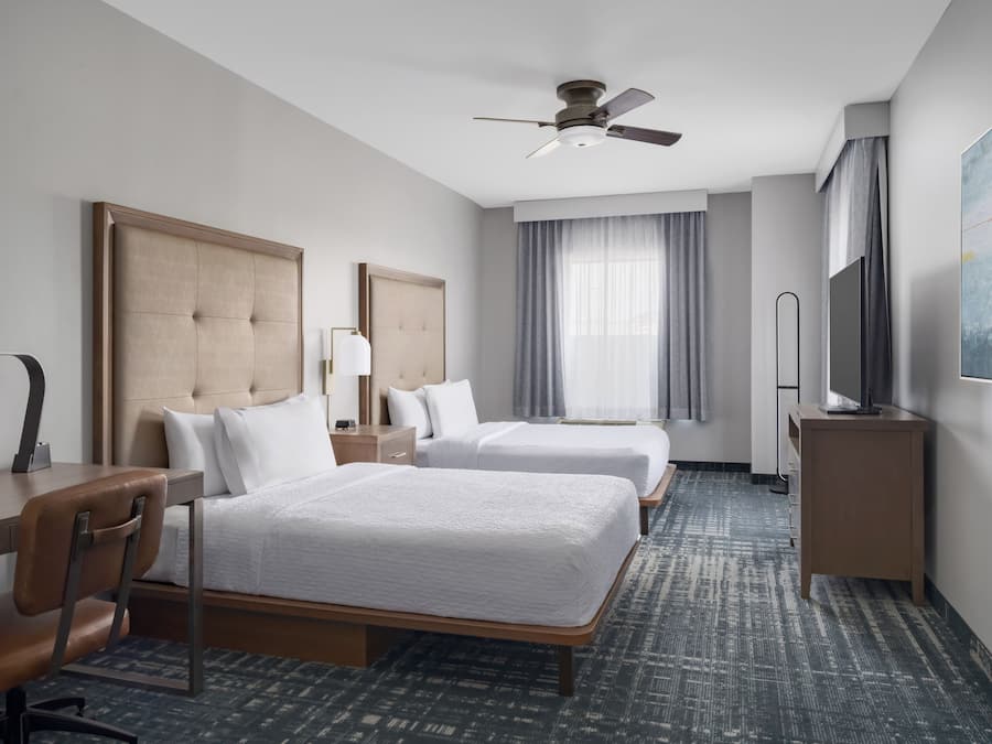 A hotel room with two beds, a desk, a chair, a ceiling fan, a TV, and a large window with curtains.
