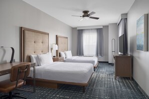 Habitación de hotel con dos camas, un escritorio, una silla, un ventilador de techo, un televisor y un amplio ventanal con cortinas.