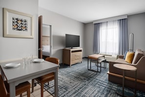 A hotel room with a dining table, chairs, a sofa, a TV, a desk, and a patterned carpet. - Homewood Suites by Hilton Midland, TX (Midland)