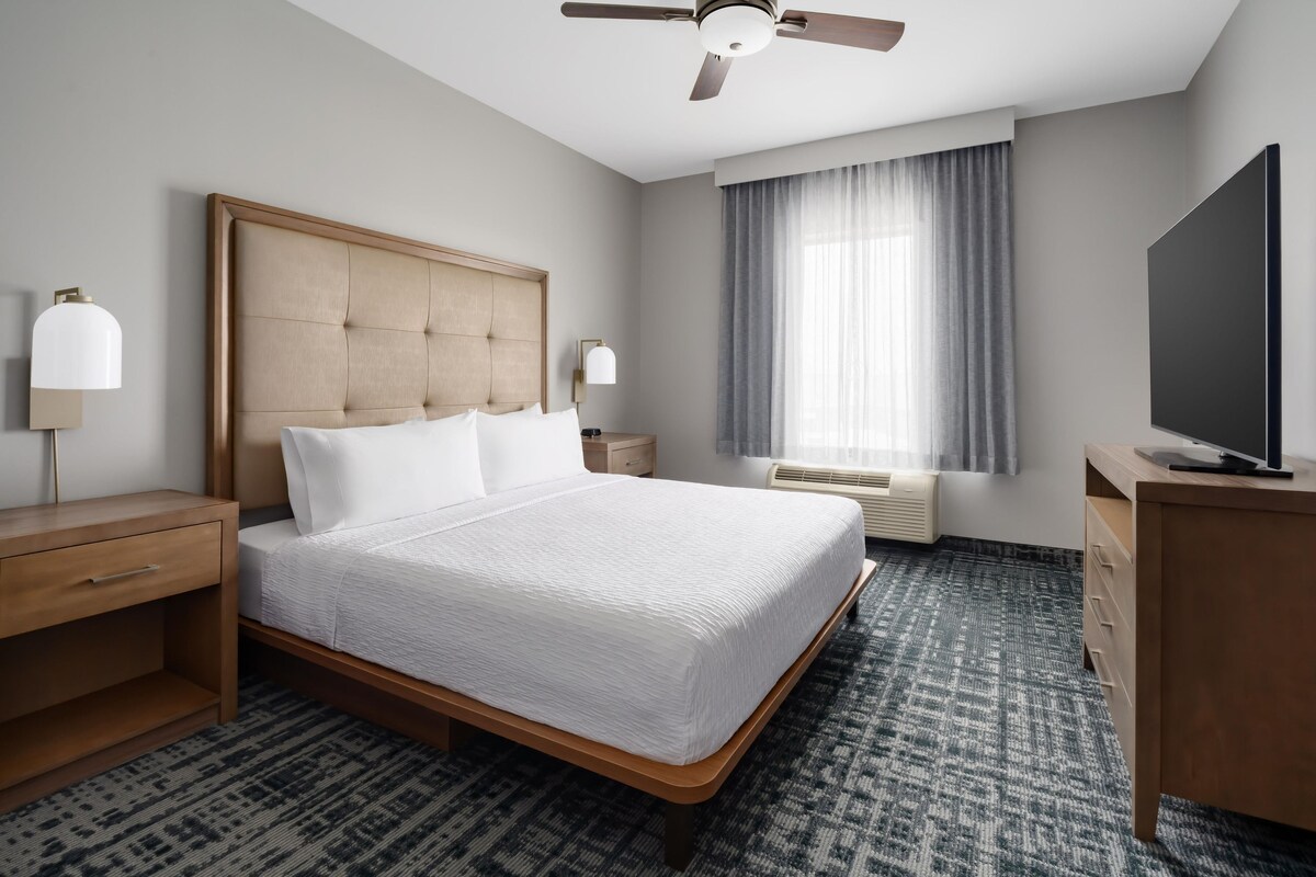 A hotel room with a large bed, a desk, a television, a ceiling fan, a window with curtains, and a wall-mounted TV.