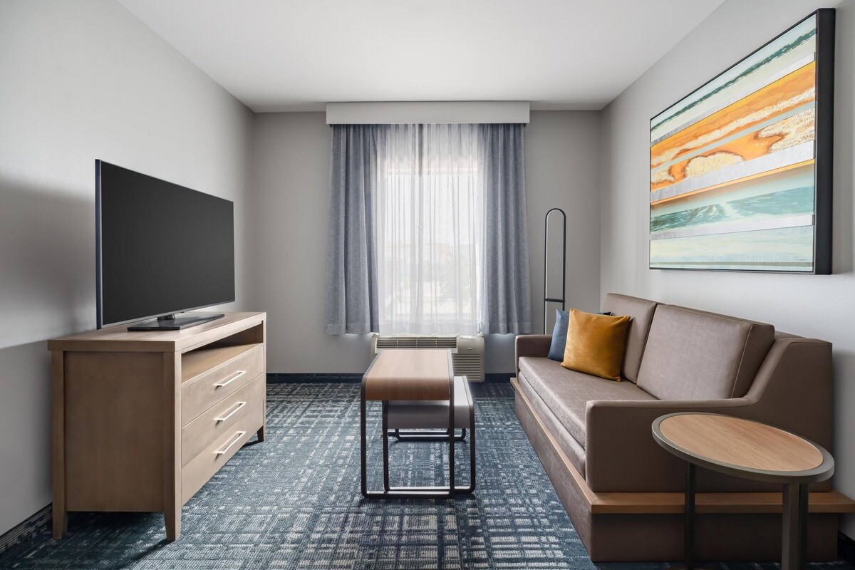 A hotel room with a flat-screen TV, a sofa, a small table, a bench, and a large window with curtains.