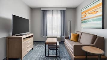 A hotel room with a flat-screen TV, a sofa, a small table, a bench, and a large window with curtains.