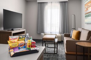 A hotel room with a flat-screen TV, a dining table with snacks, a sofa, a small table, and a window with curtains. - Homewood Suites by Hilton Midland, TX (Midland)