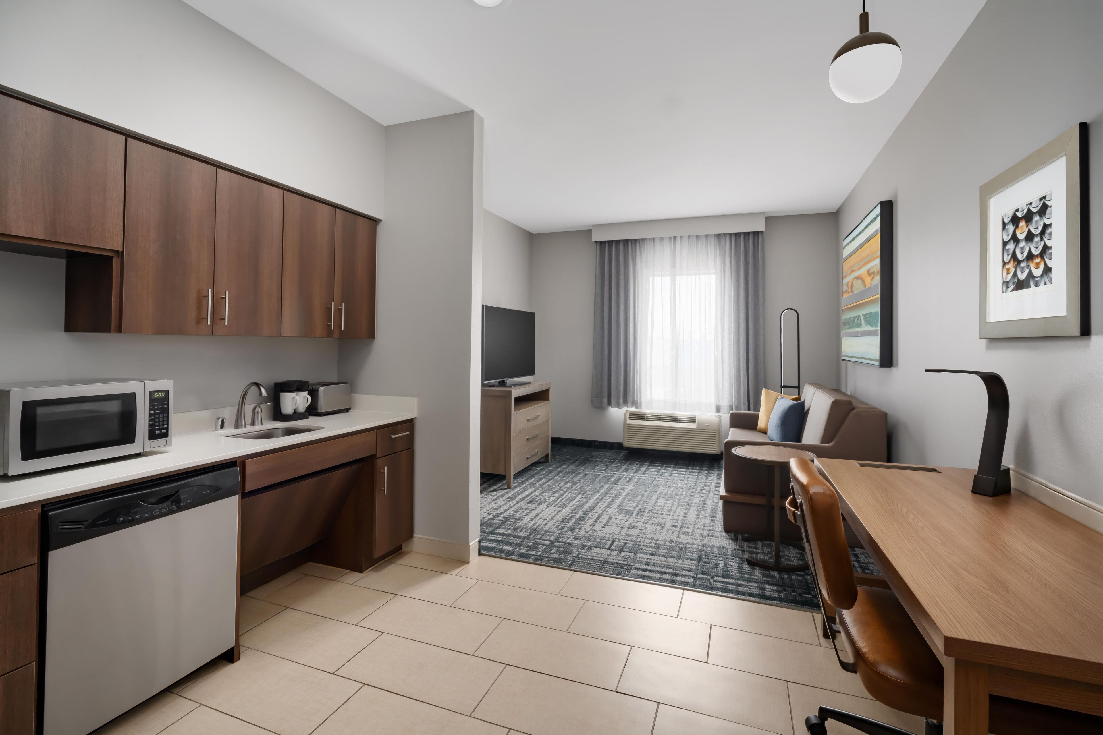 A modern kitchen with a dishwasher, microwave, and a flat-screen TV. There's a dining area with a wooden table and chairs, and a living space with a sofa and a TV.