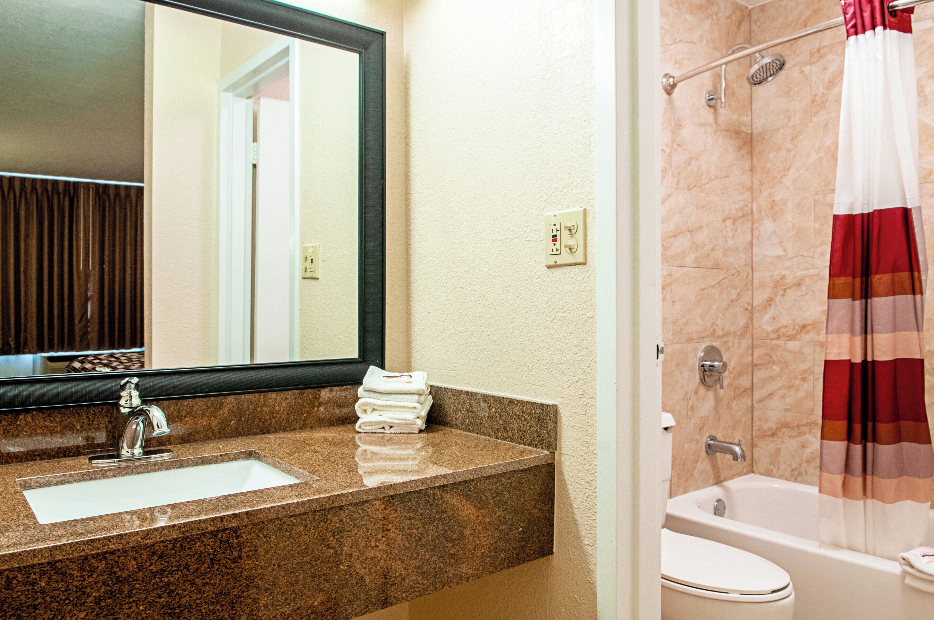 A bathroom with a granite countertop sink, a mirror, a shower with a striped curtain, and a bathtub.