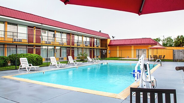 A swimming pool area with lounge chairs and umbrellas, a building with multiple windows and balconies, and a fenced yard. - Red Roof Inn Slidell  (Slidell)