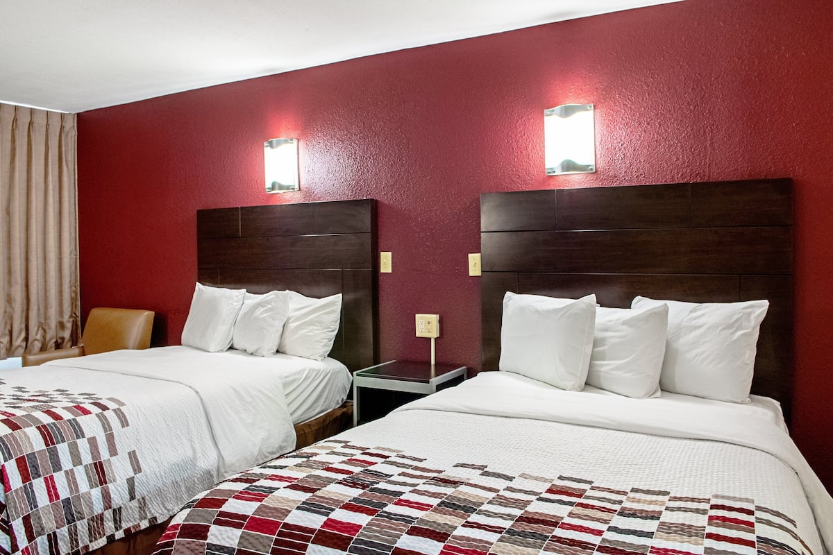 Two twin beds with white linens, a red wall, and a small bedside table with a lamp.