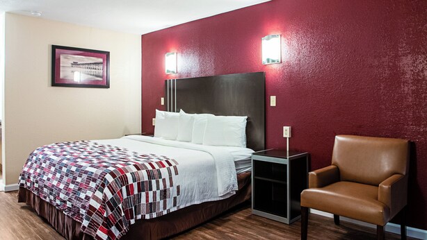 A hotel room with a bed, a chair, a nightstand, and a wall-mounted light fixture. - Red Roof Inn Slidell  (Slidell)