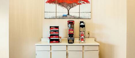 Un área de recepción con una máquina de café, un dispensador de bebidas y una pila de cajas rojas sobre un mostrador.