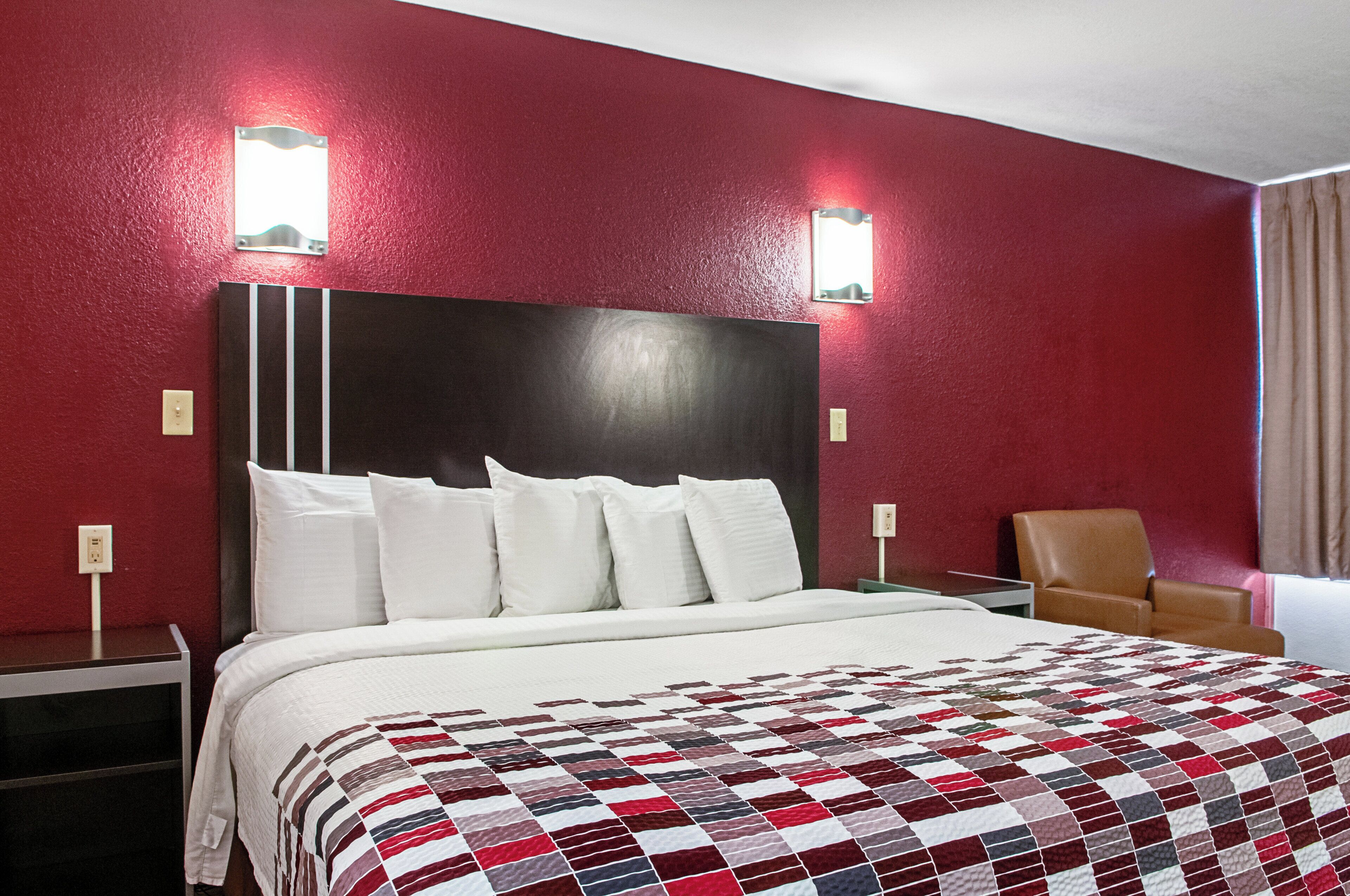Una habitación de hotel con una cama grande, cabecera, dos luces fijadas a la pared, una silla y un ventanal con cortinas.