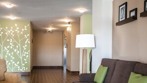 A modern living room with a brown sofa, a round coffee table, and a green cushion. There is a wall with a white branch design and a floor lamp. - Sleep Inn & Suites Gulfport (Gulfport)