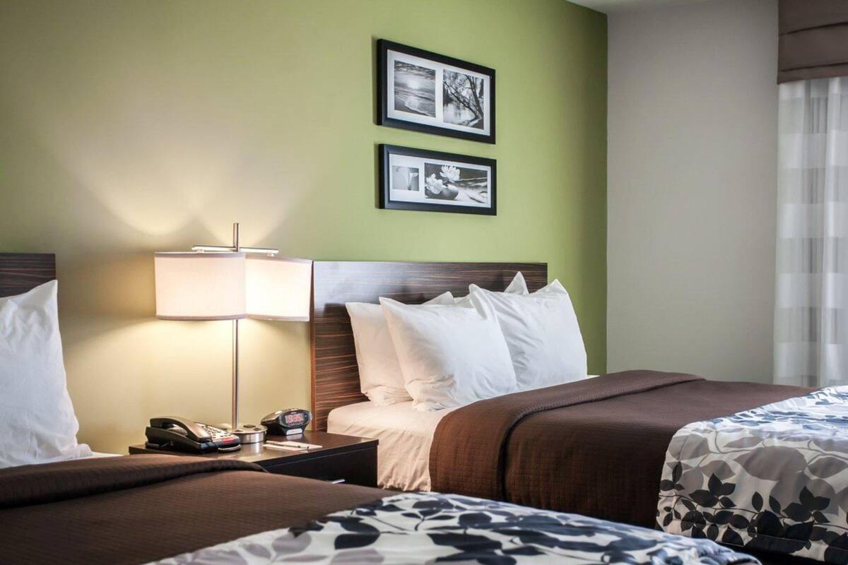 A hotel room with two beds, a nightstand with a lamp, a phone, and a clock. There are two framed pictures on the wall.