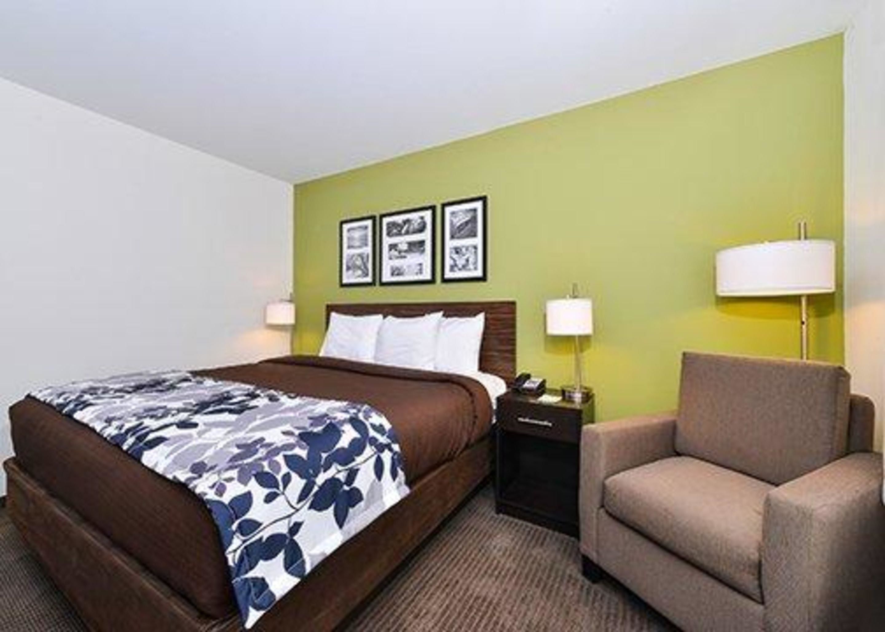 A hotel room with a bed, a chair, a nightstand, and a wall with framed pictures.