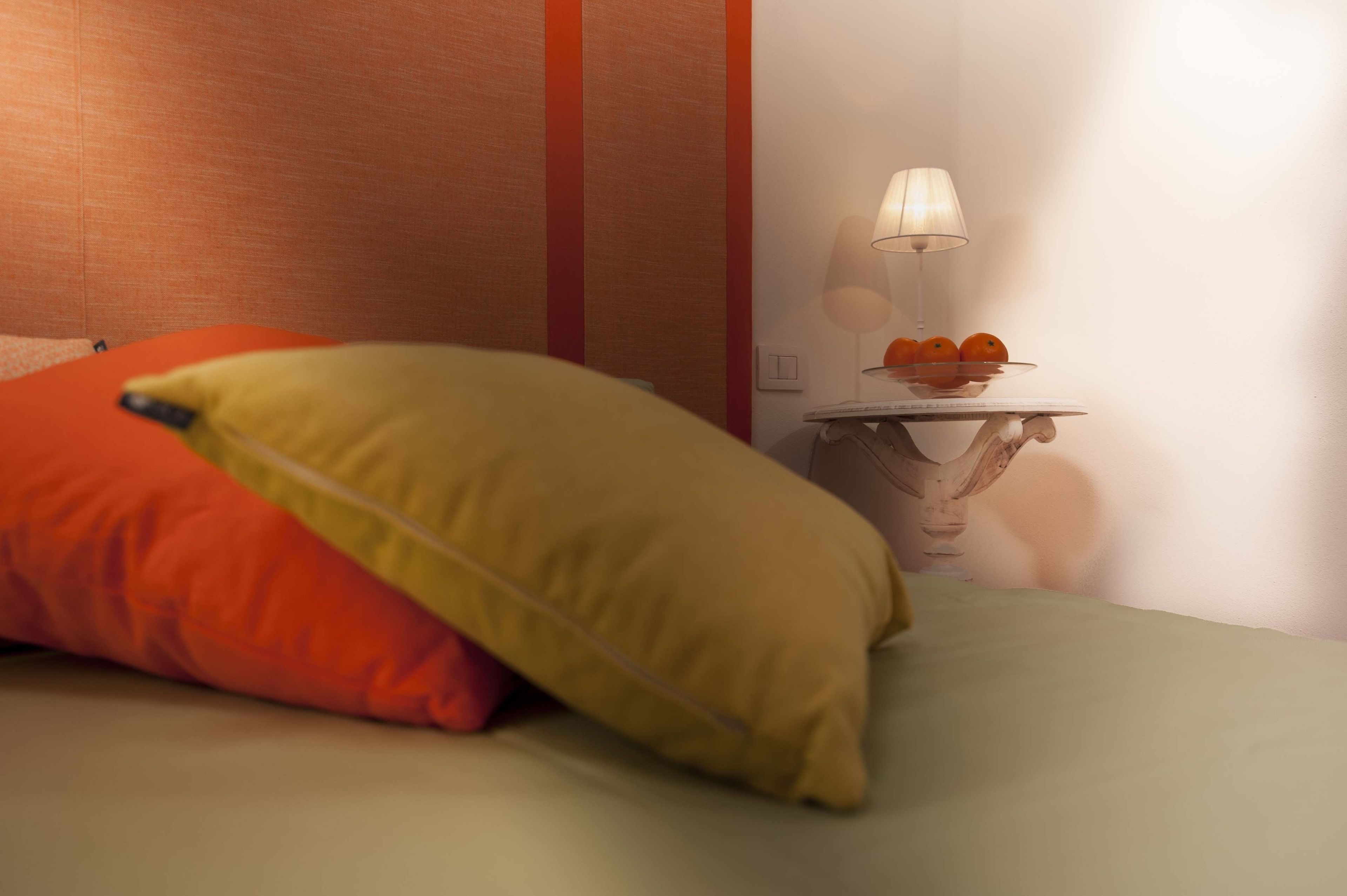Ein Bett mit orangefarbenen und gelben Kissen, ein Nachttisch mit Lampe sowie eine Wand mit rotem Streifen.
