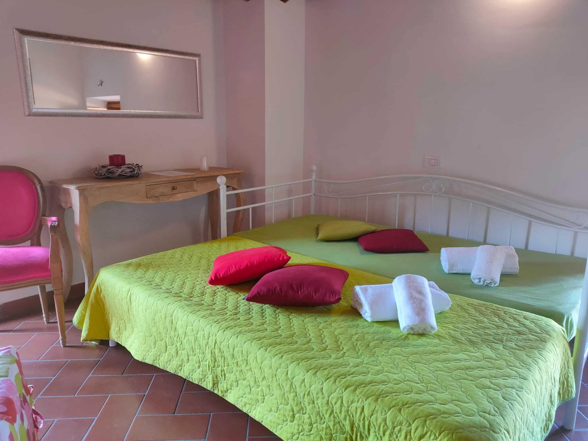 A bedroom with two beds, each with green bedding and multiple pillows. There is a wooden desk with a chair and a mirror on the wall.