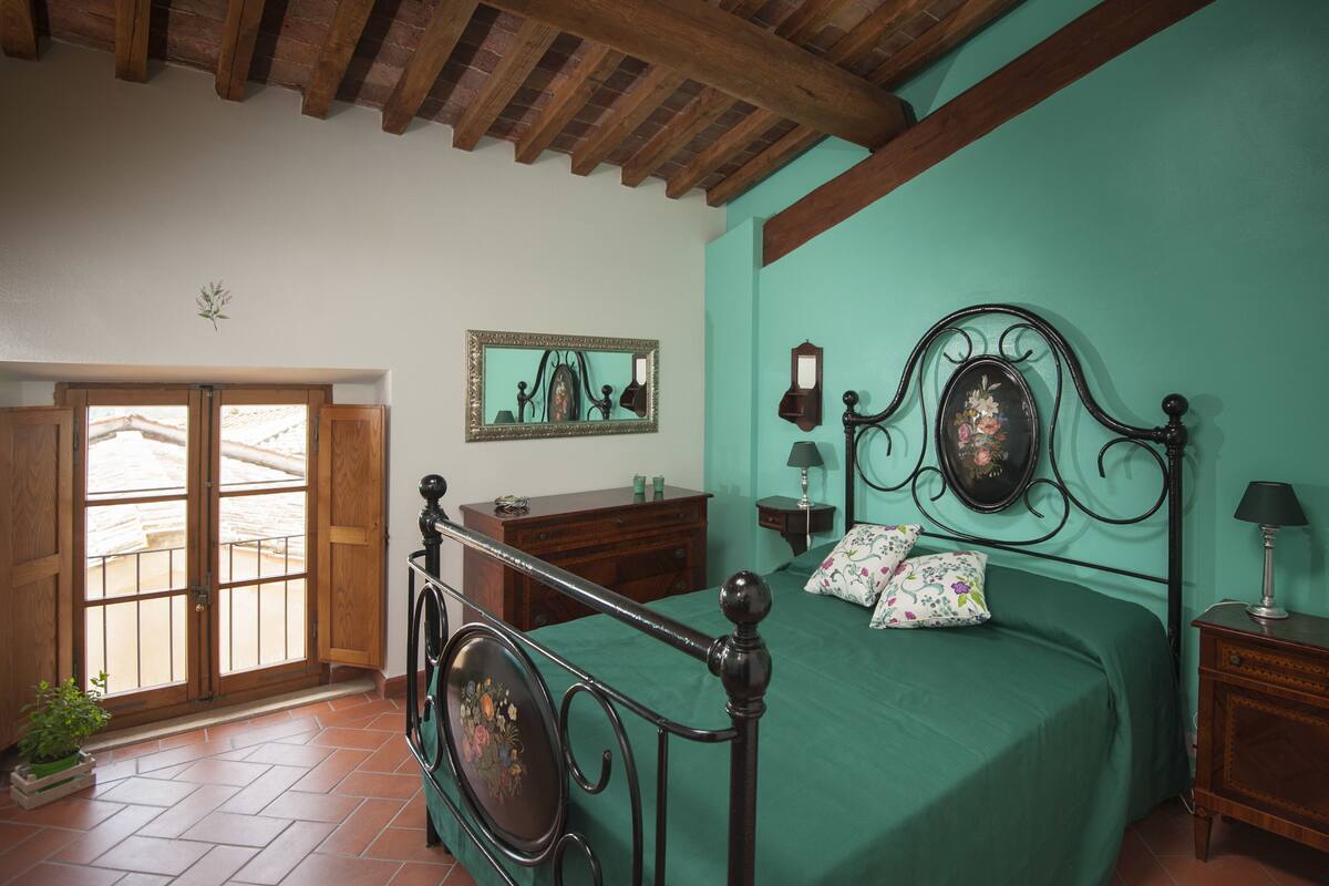 A bedroom with a wooden bed frame, a green bedspread, a wooden dresser, a mirror, and a window with a view of a balcony.