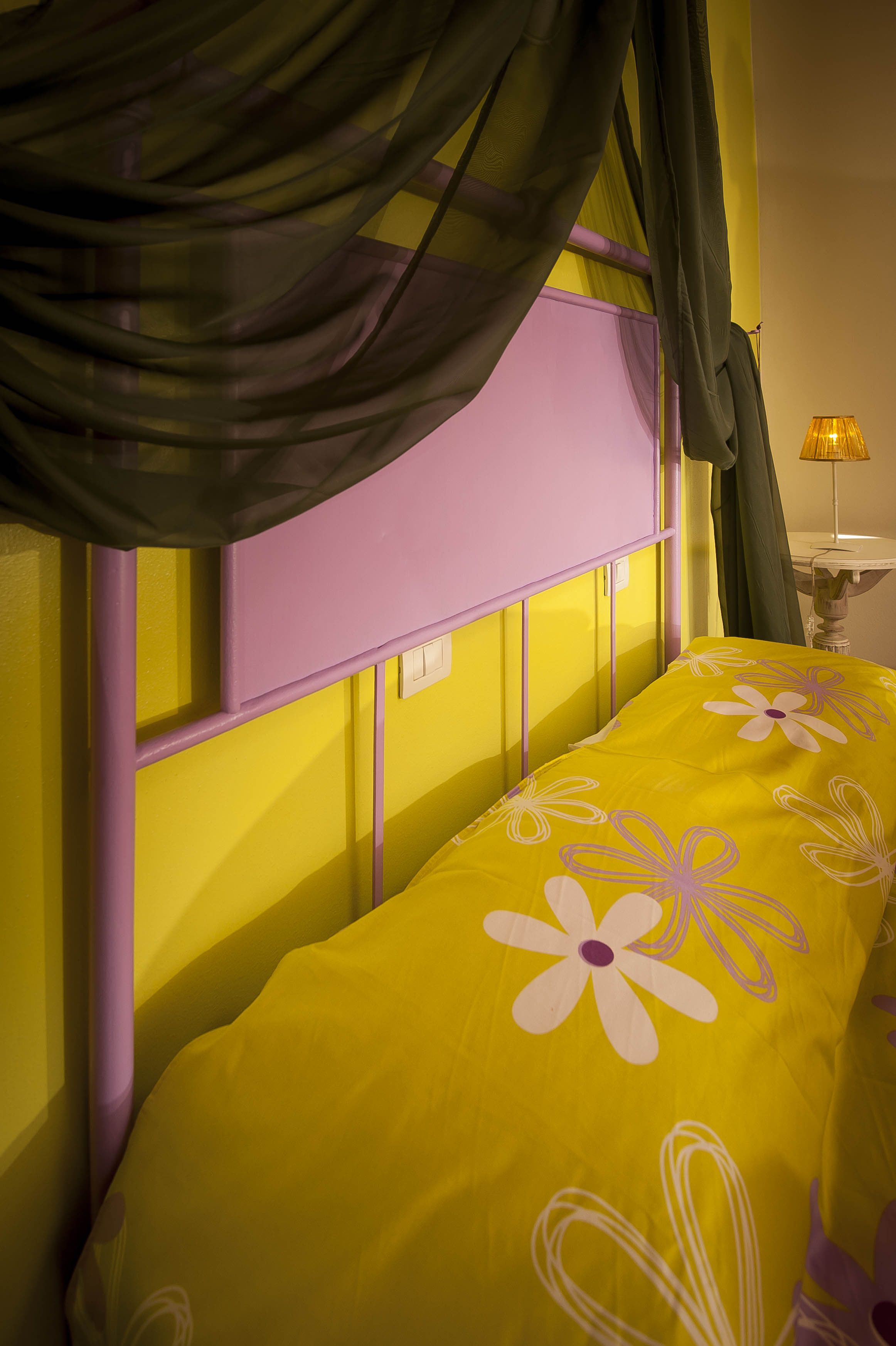 Una cama con colcha amarilla estampada con flores, cabecera morada y cortinas de encaje.