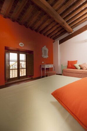 Un dormitorio con techo de madera, pared naranja, cama con ropa de cama naranja, cabecera verde, mesita de noche pequeña y ventana con vistas a un balcón.