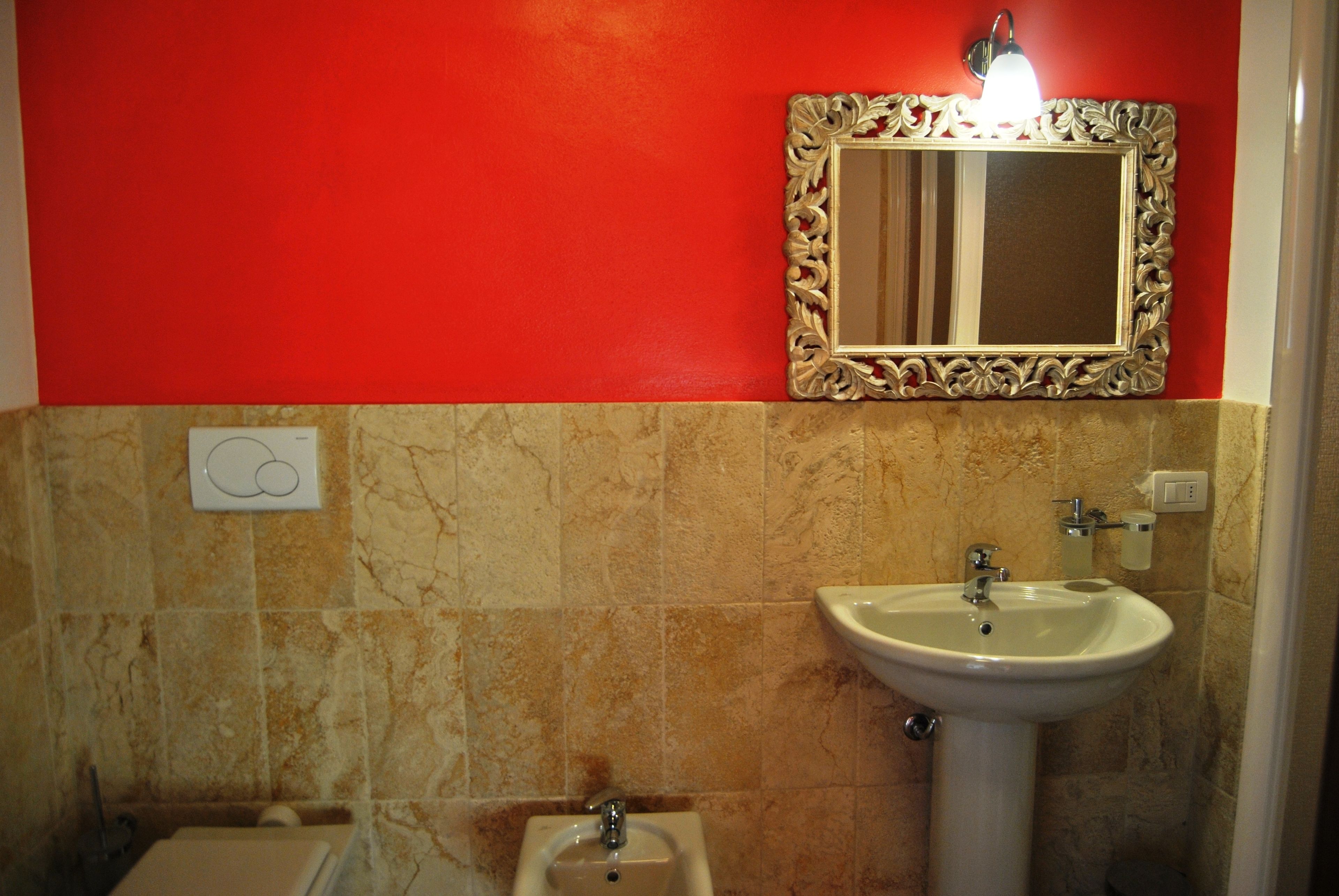 Ein Badezimmer mit roter Wand, Marmorboden, einem Standwaschbecken, einem an der Wand befestigten Spiegel und einer Toilette.