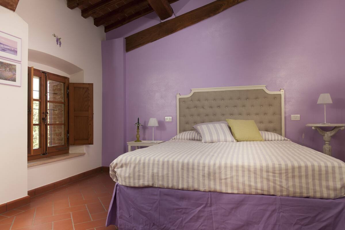 A bedroom with a bed, wooden ceiling beams, a window with shutters, a nightstand, and a lamp.