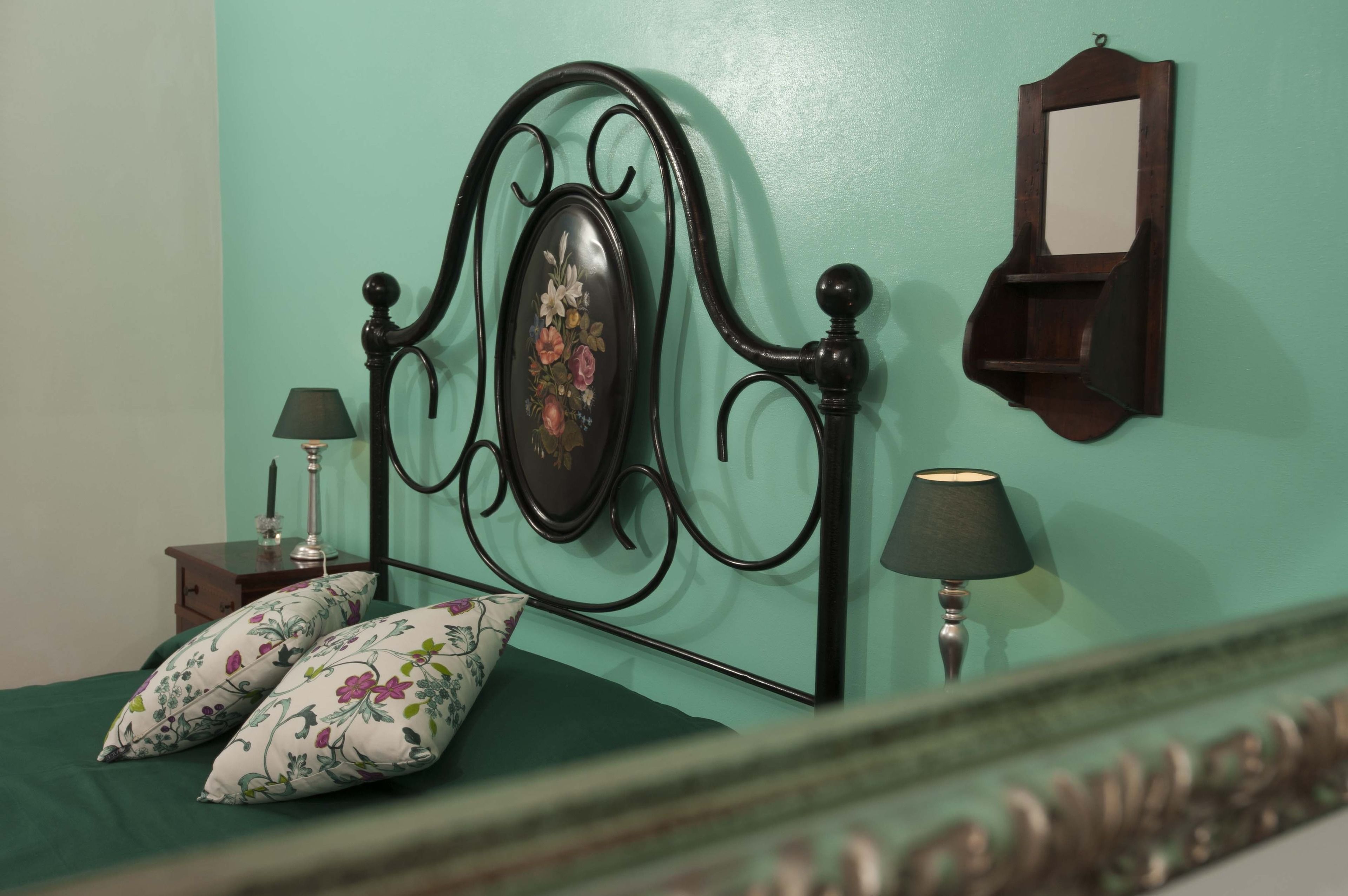 Un lit avec une tête de lit à motifs floraux, deux oreillers, une table de chevet avec une lampe et un miroir fixé au mur.