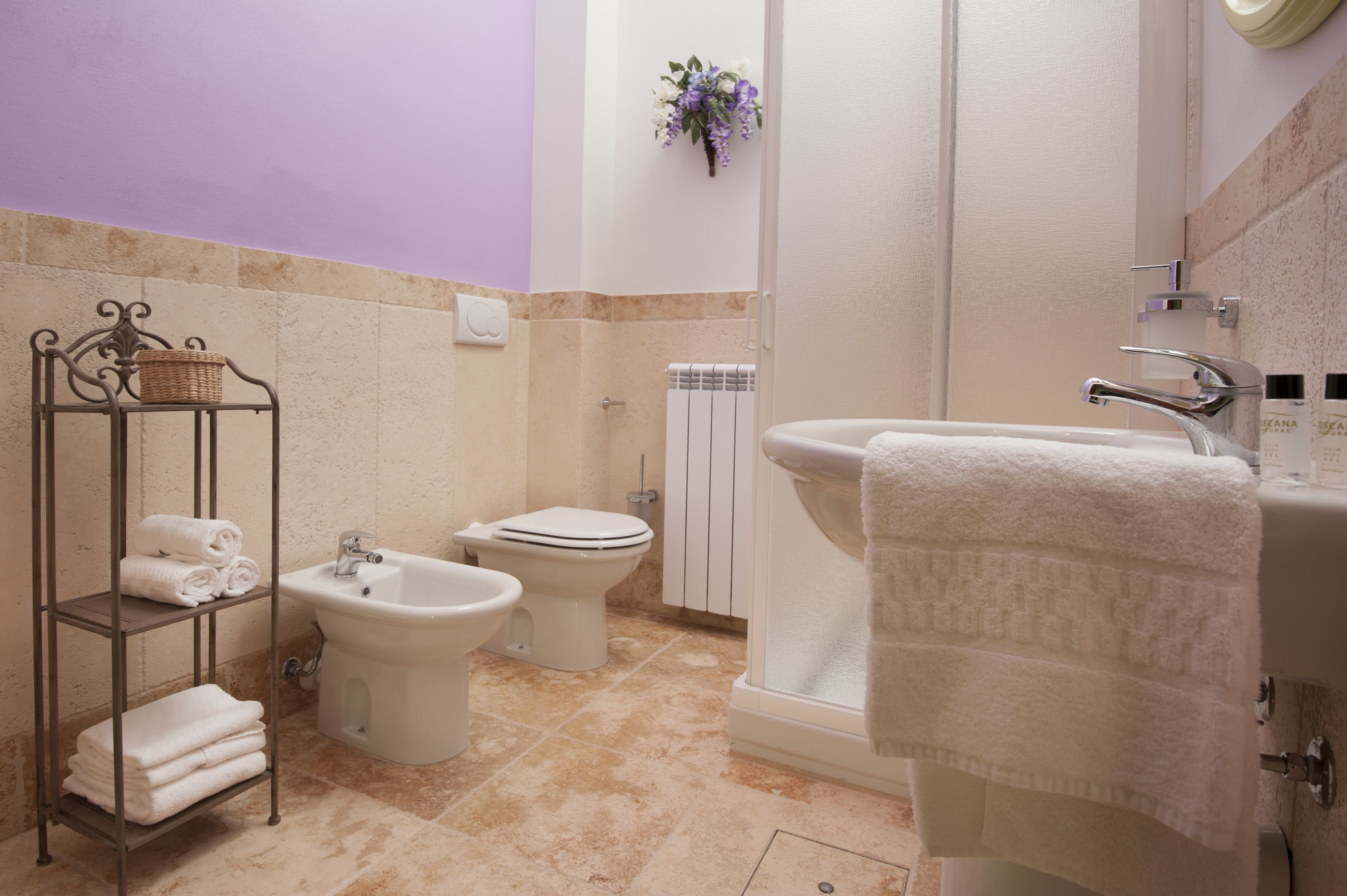 Un baño con inodoro, bidé y lavabo. Hay un perchero con toallas dobladas y una cesta.