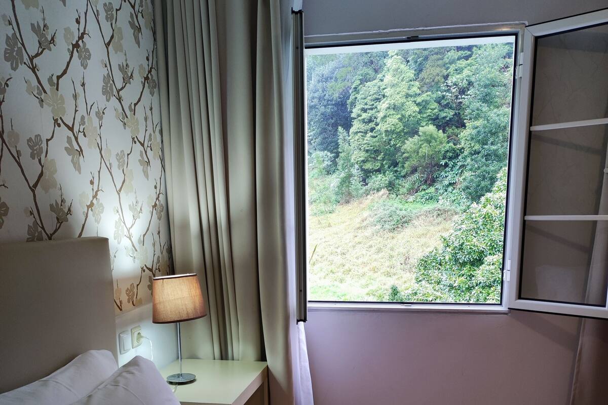 Makuuhuoneessa on sänky, yöpöytä, lamppu, ikkuna, josta on näkymä vehreään maisemaan, ja seinä, jossa on kukallinen tapetti.