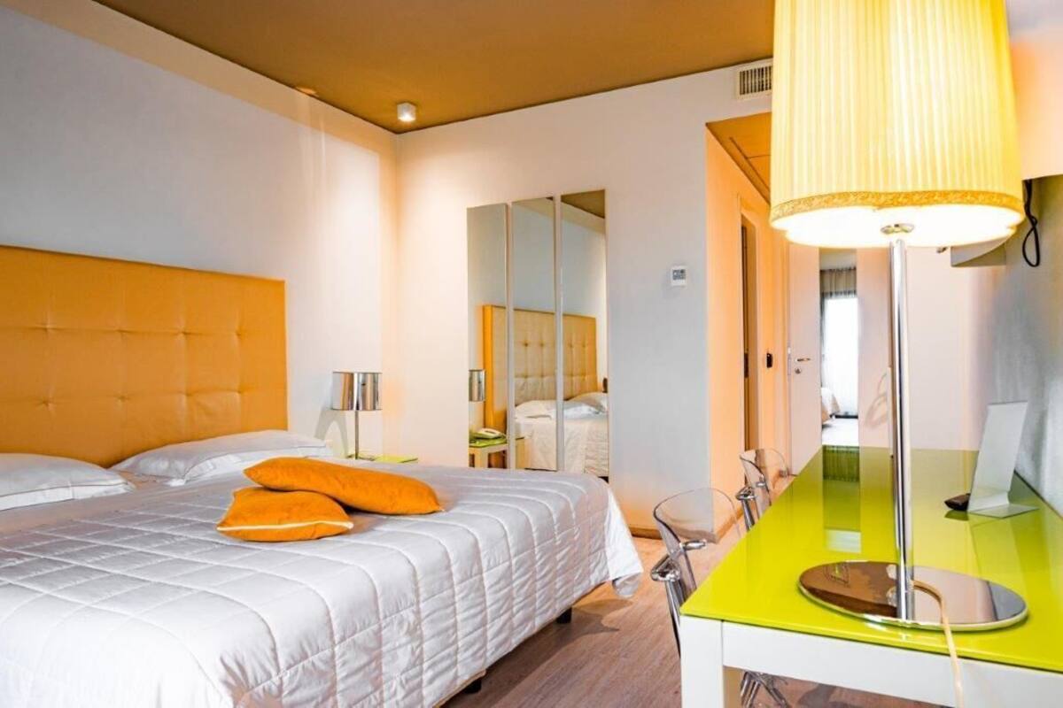 Quarto de hotel com uma cama grande, uma mesa de cabeceira, um espelho e uma luminária clara.