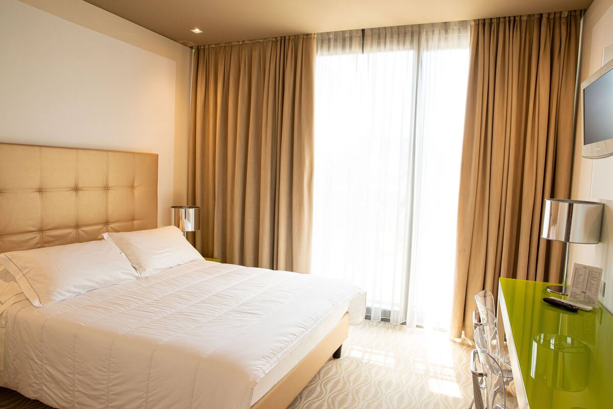 Quarto de hotel com uma cama grande, uma escrivaninha verde e uma televisão na parede.