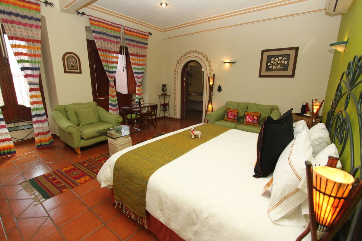 A hotel room with a large bed, green sofa, and a small table. There is a door with curtains and a framed picture on the wall.