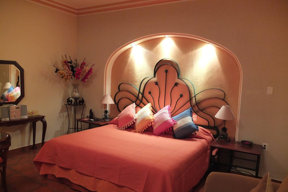 A bedroom with a bed, two bedside tables, a chair, a mirror, and a vase with flowers.