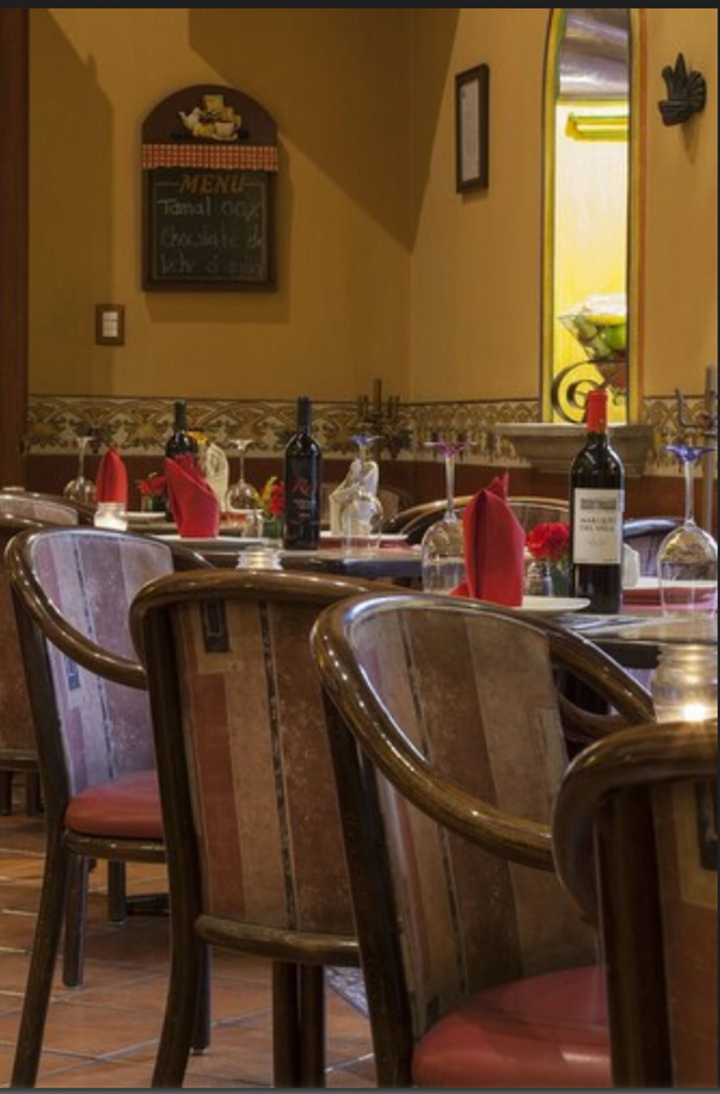 Interno di un ristorante con sedie di legno, un menu appeso al muro e bottiglie di vino sul tavolo.