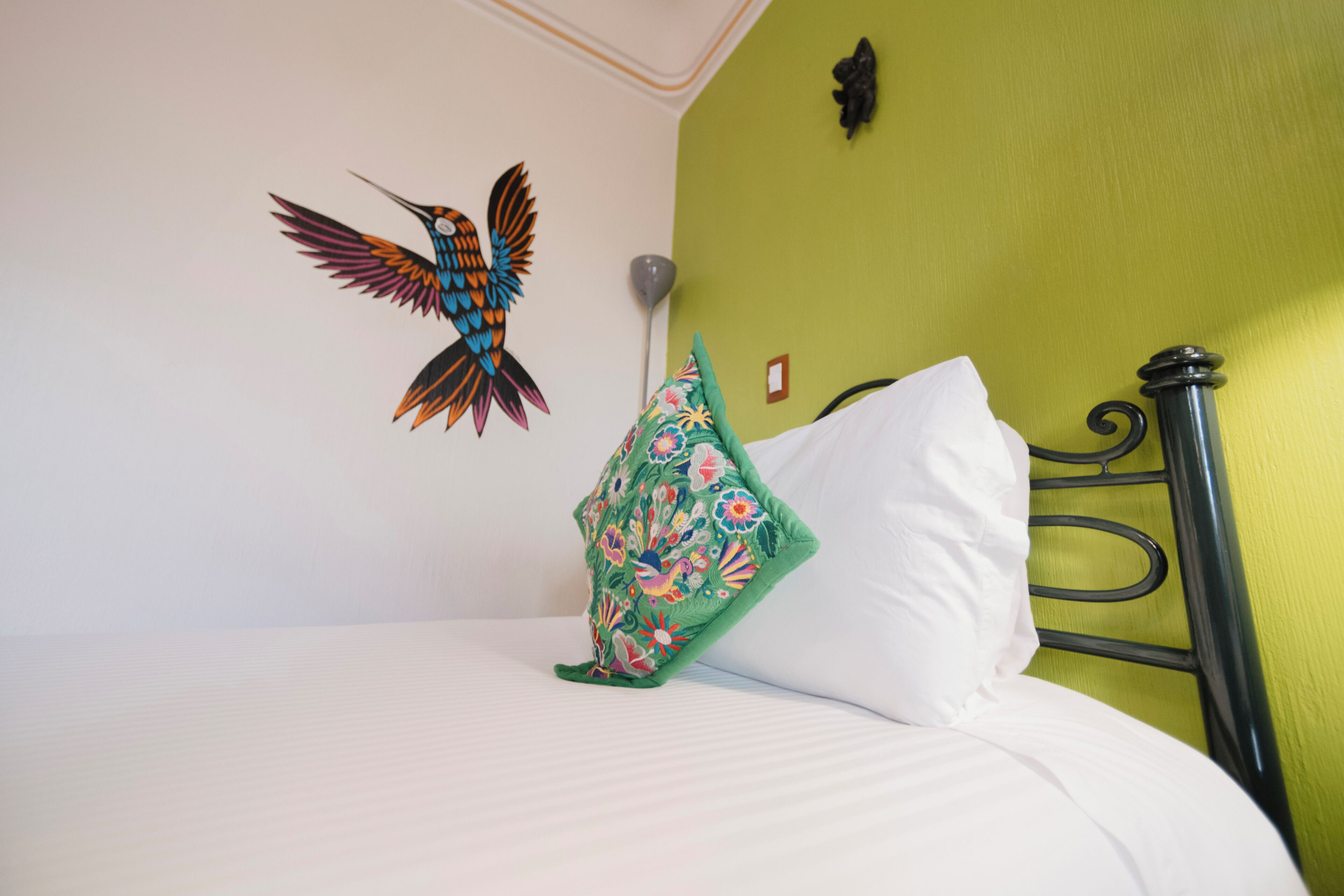 Una cama con ropa de cama blanca, un cojín verde con motivos florales y un adorno de pájaro colgado en la pared.