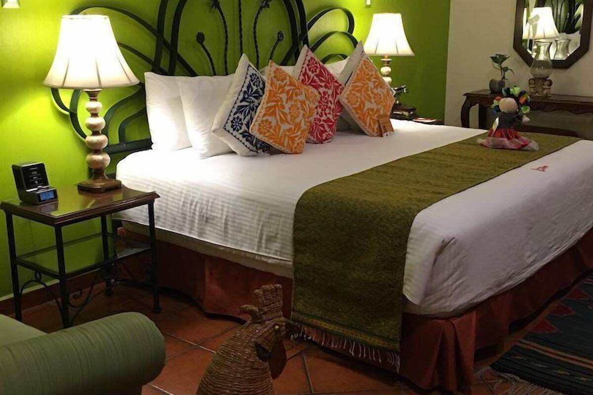 A hotel room with a bed, two bedside lamps, a bedside table with a phone, a green wall, a mirror, and a small plant.