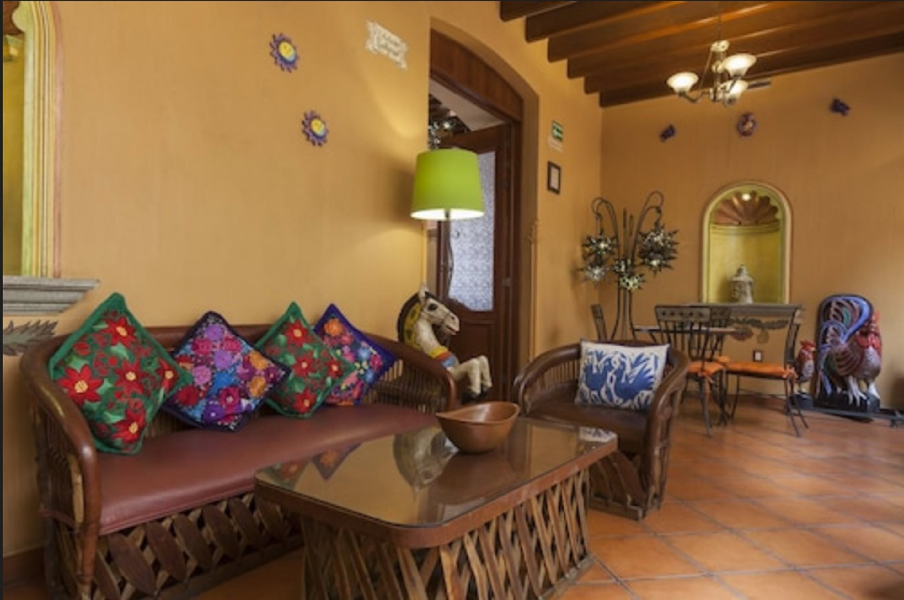 A cozy living room with a wooden sofa adorned with colorful cushions, a glass coffee table, and a rustic dining area with a wooden table and chairs.