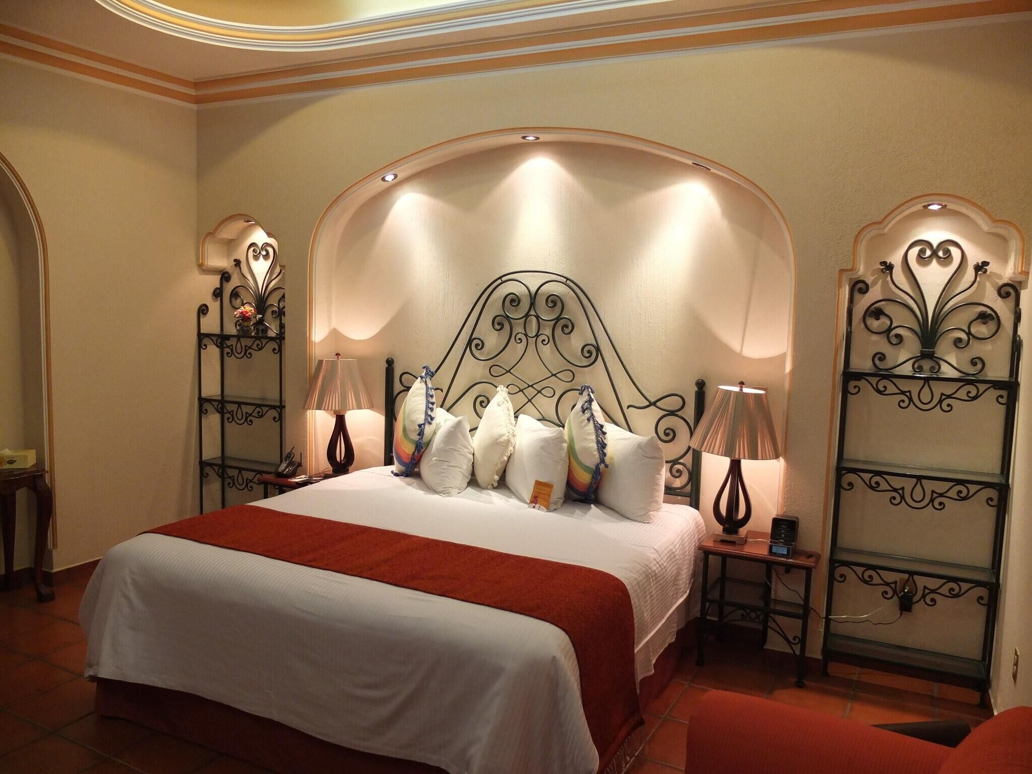 A hotel room with a large bed, two bedside tables, a red chair, and decorative metal railings.