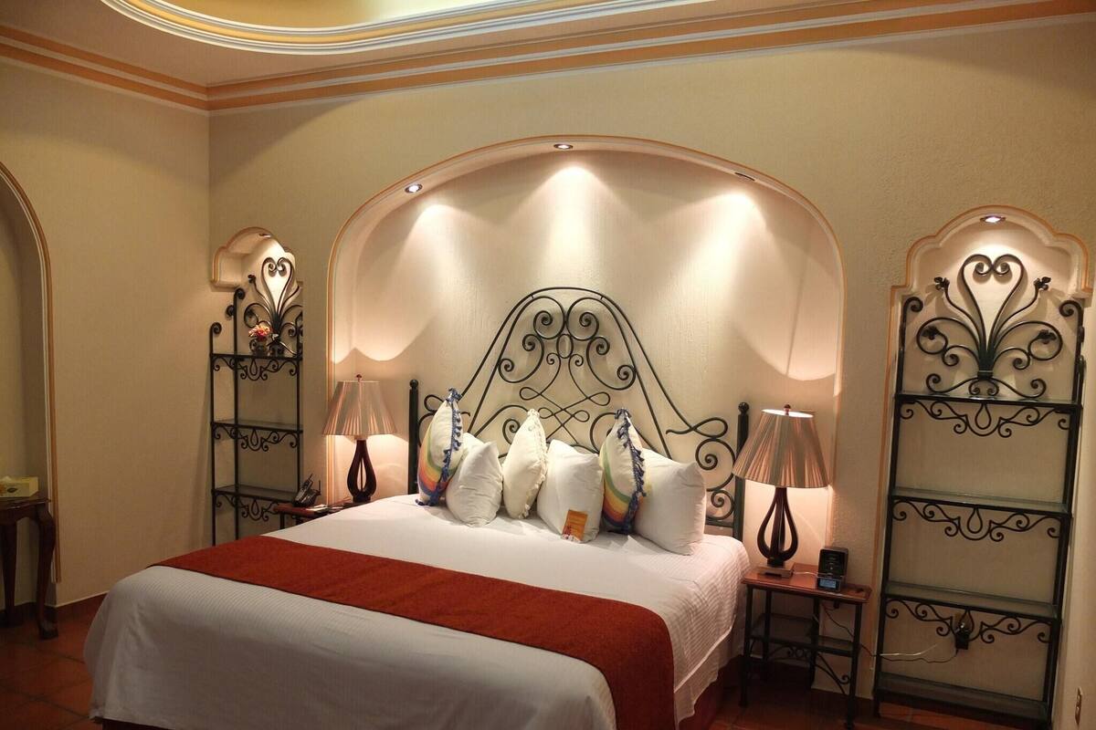 A hotel room with a large bed, two bedside tables, a red chair, and decorative metal railings.