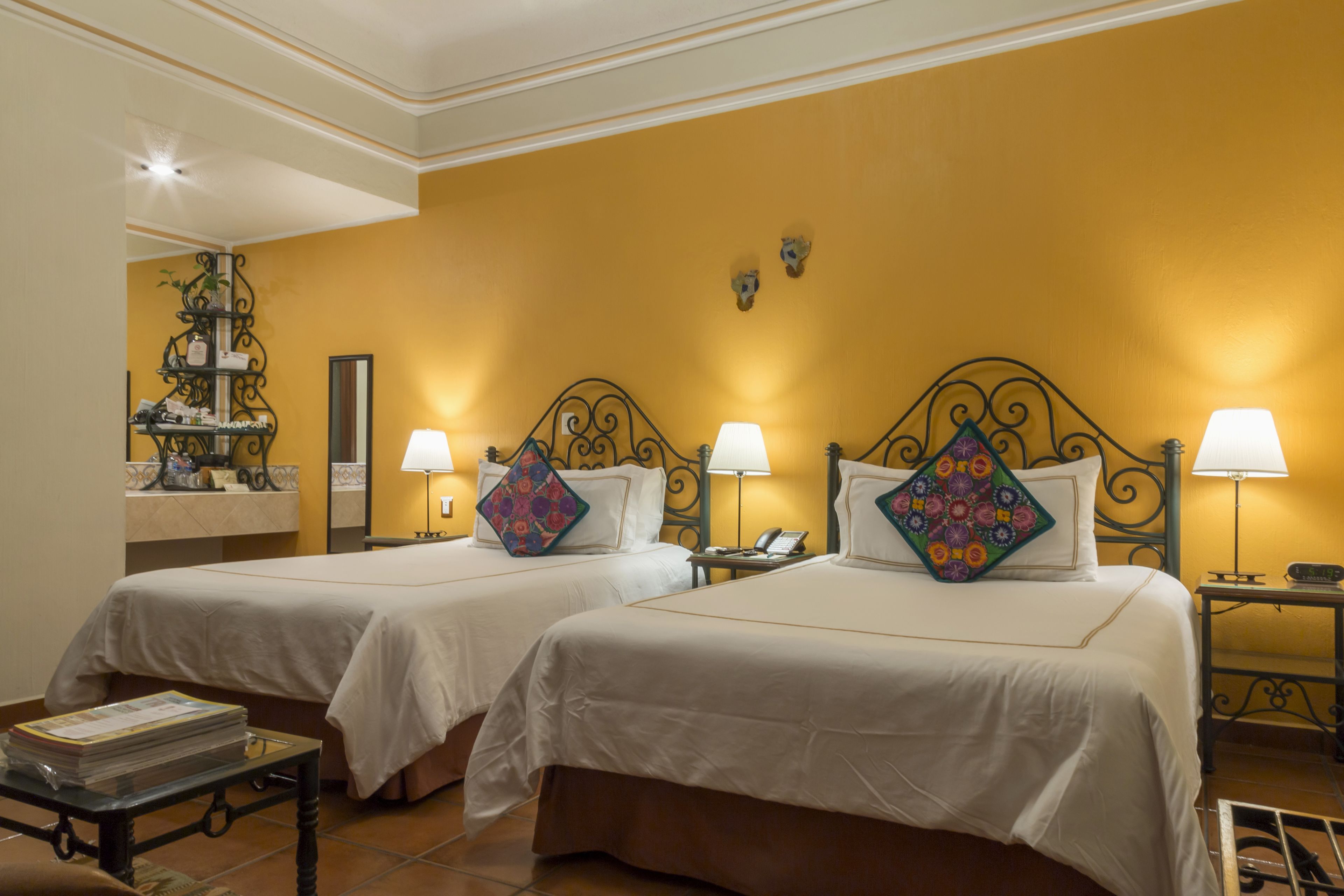 Une chambre d’hôtel avec deux lits, chacun recouvert d’une courtepointe blanche et agrémenté de coussins décoratifs. On y trouve des tables de chevet avec des lampes, une petite table sur laquelle est posée une pile de magazines, ainsi qu’une étagère murale décorative.