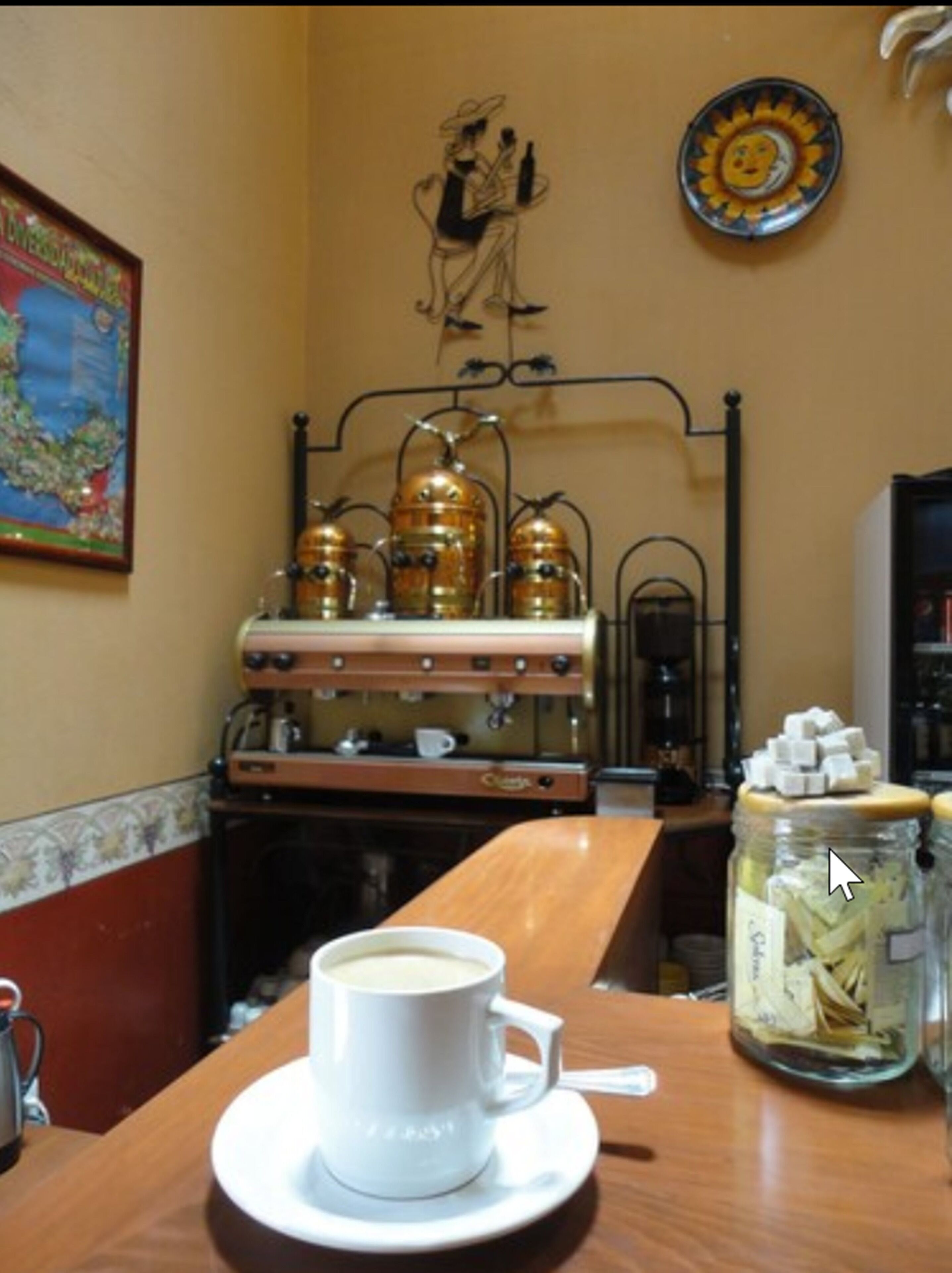 Interior de una cafetería con una taza de café sobre un platito, una máquina de café, un estante metálico decorativo y un tarro de azúcar en la barra.