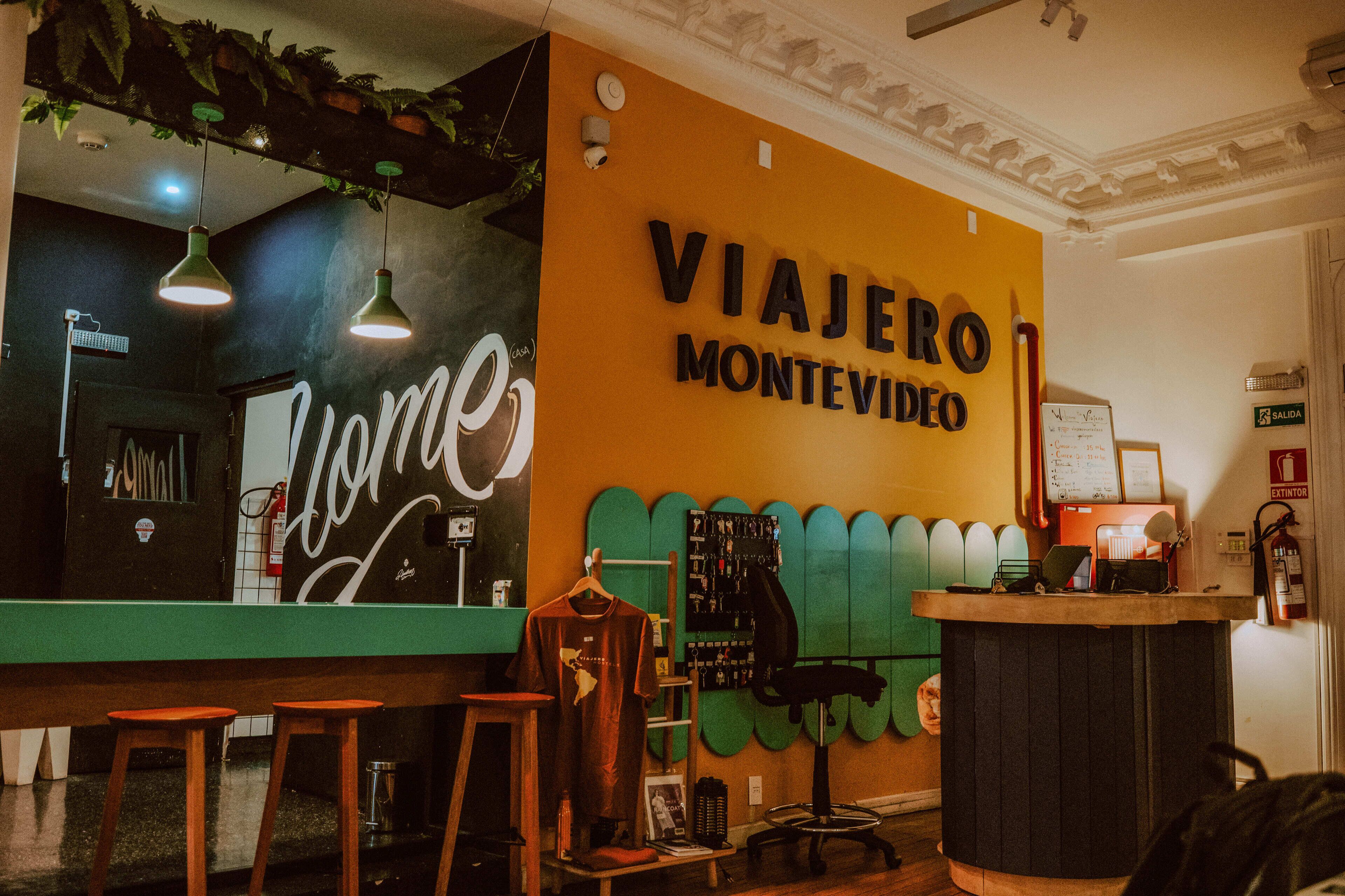 Un área de recepción con un gran letrero que dice "VIAJERO MONTEVIDEO", una recepción, una silla y una pared decorada con motivos de hojas verdes.