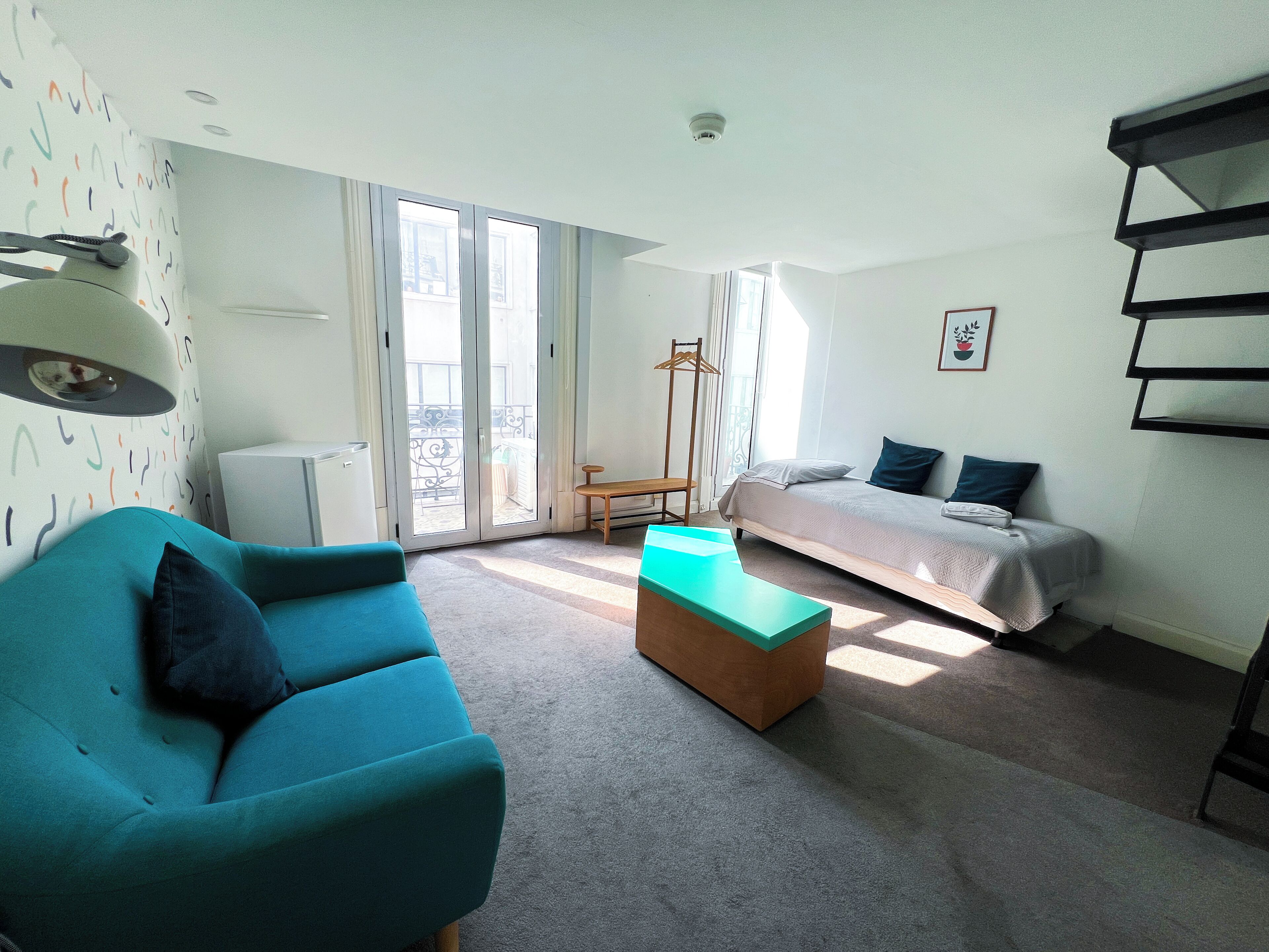 A modern room with a teal sofa, a bed with two pillows, a small wooden table, and a large window with a balcony.