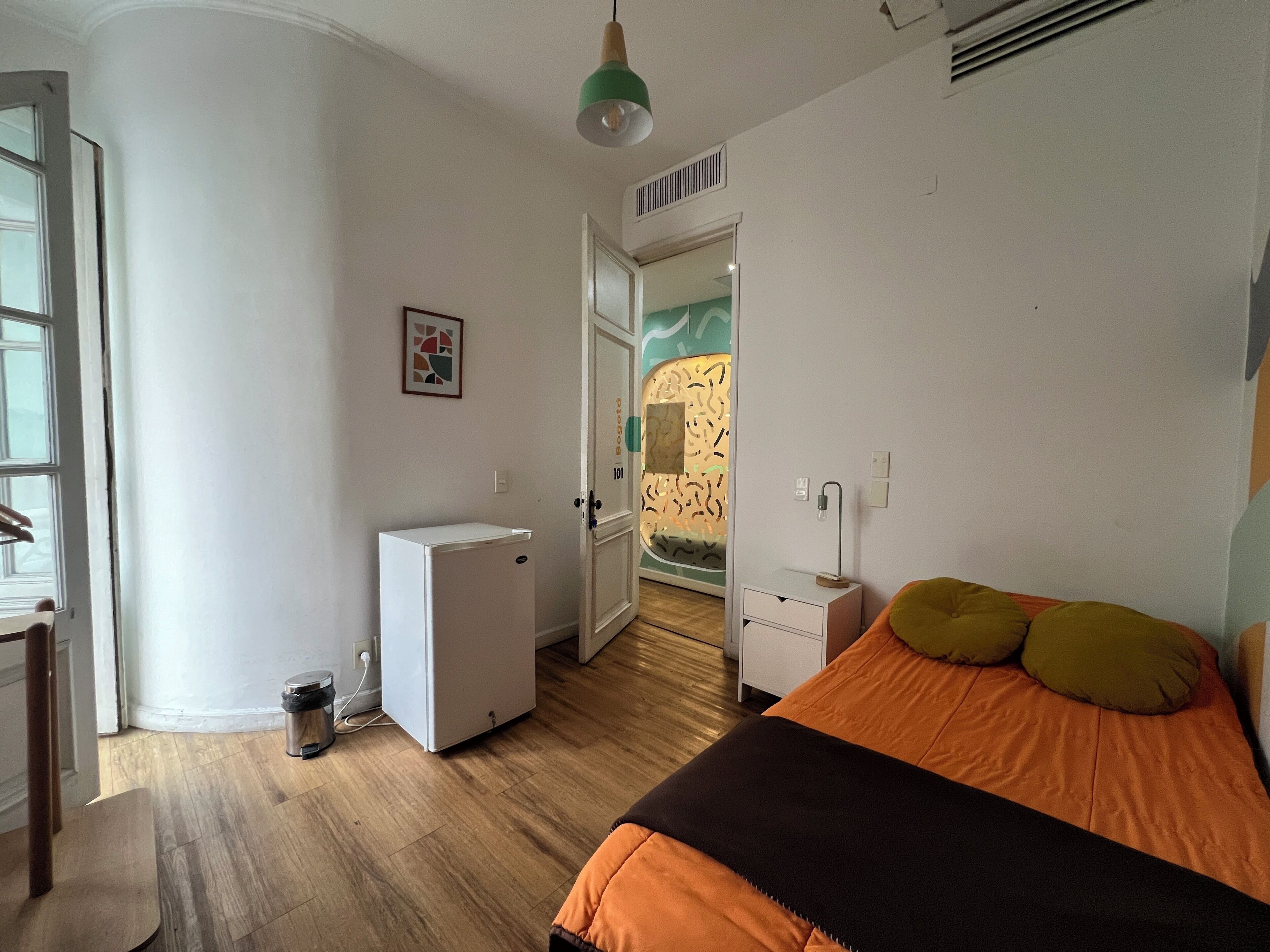 Un dormitorio con una cama, un escritorio, una silla, un refrigerador blanco pequeño y una puerta que da a otra habitación.