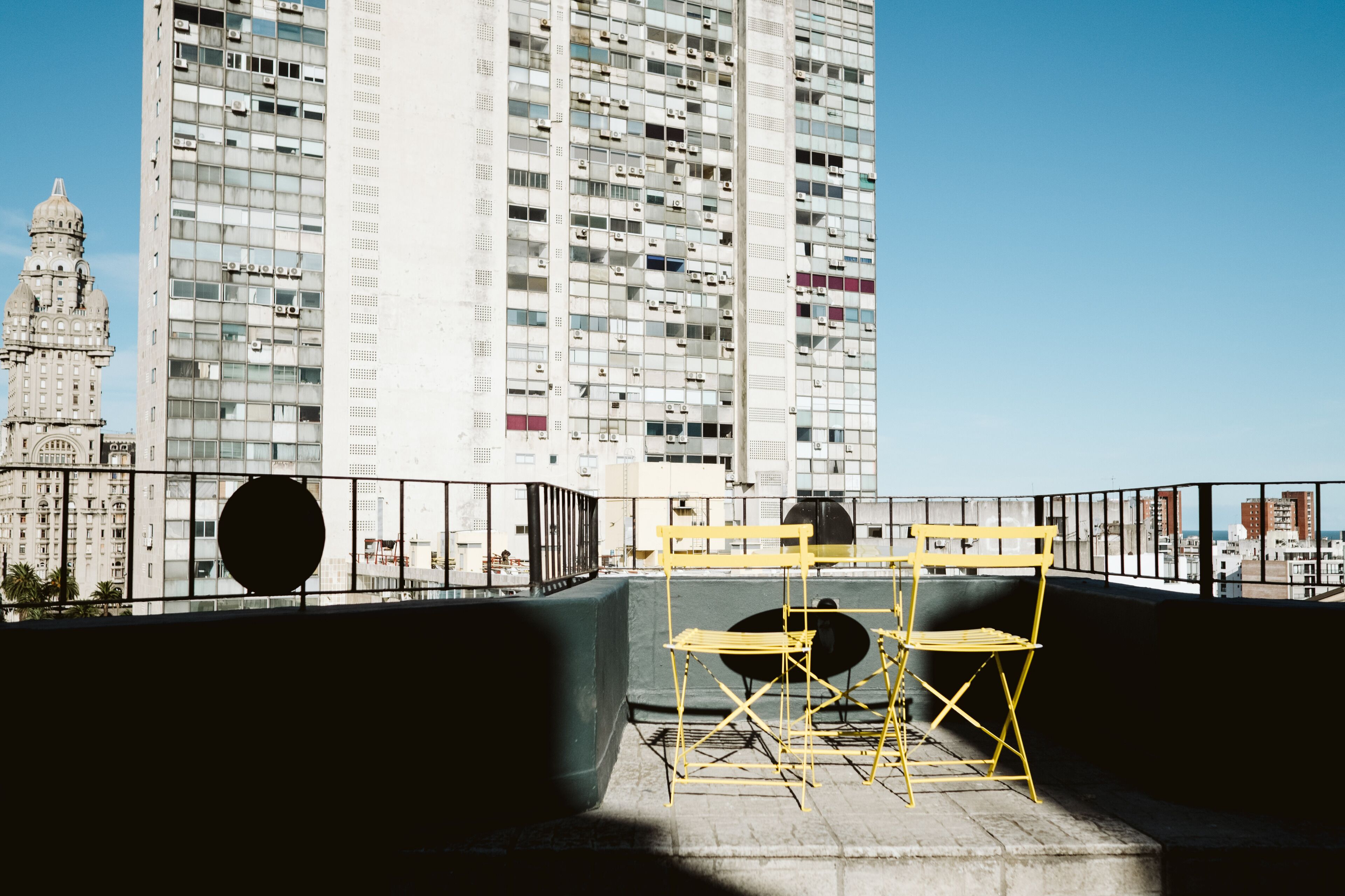 Une terrasse sur le toit, avec une chaise pliante jaune et un objet circulaire noir, d’où l’on peut admirer une vue sur la ville, avec ses bâtiments modernes et historiques.
