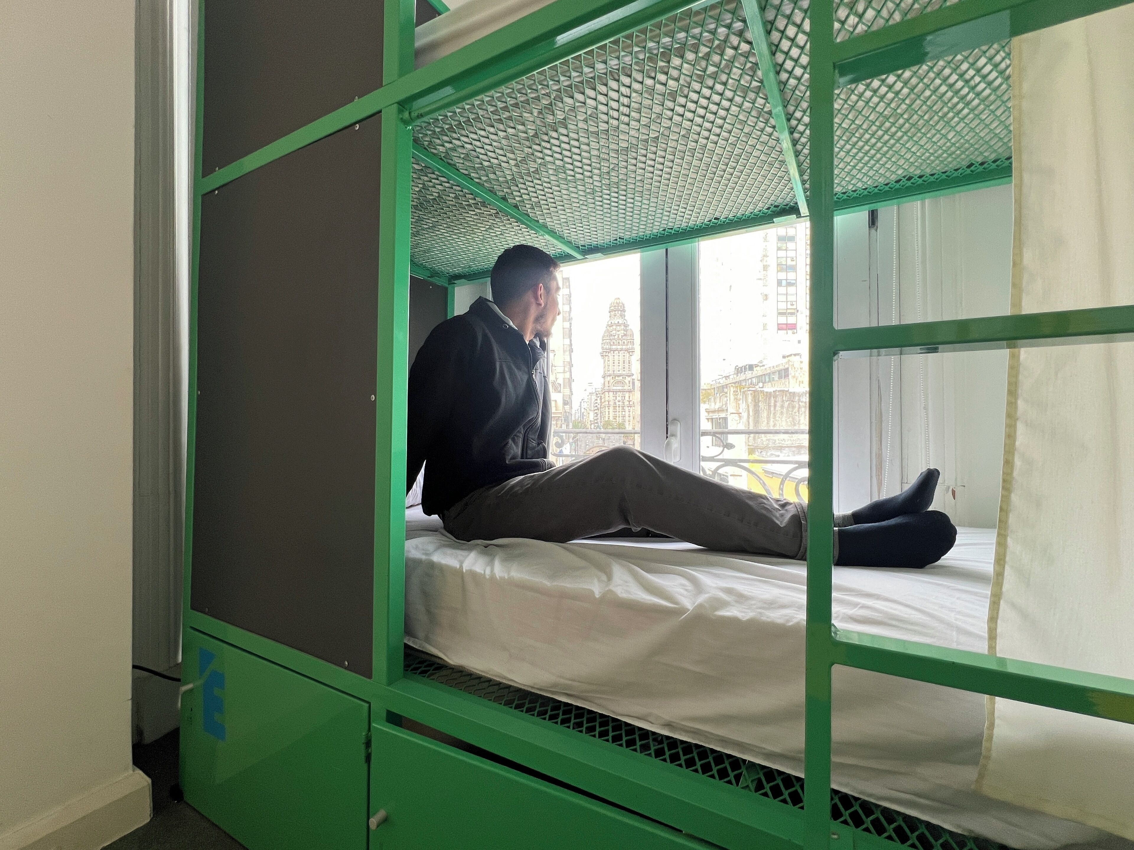 A bunk bed with a person sitting on the lower bunk, a window with a view of buildings, and a green metal frame.