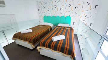 Habitación de hotel con dos camas, cada una con ropa de cama y toallas a rayas. La habitación cuenta con un panel de vidrio, un cabecero verde azulado y un papel tapiz estampado.
